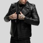 Seitlich posierender Mann in schwarzer Leder-Bikerjacke mit Nietenbesatz, Reißverschlussdetails und Tattoos an Händen und Hals